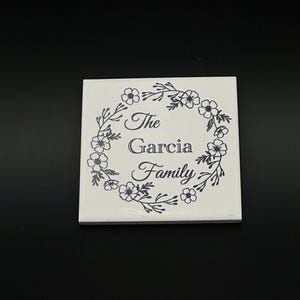 May include: A square, white ceramic tile featuring a black floral wreath design. The words "The Garcia Family" are centered within the wreath. The tile is set against a black background.