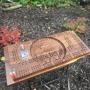 Può includere: Un tabellone di cribbage in legno marrone scuro con un'aquila incisa, stelle e foglie di alloro. Il tabellone ha molti fori per i pioli. Un mazzo di carte Bicycle Jumbo e pioli colorati sono sul tabellone.