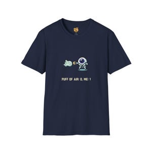 May include: Navy blue t-shirt featuring a cartoon eye character with a cloud and the text "PUFF OF AIR: 0, ME: 1". The shirt has a crew neck and short sleeves. A small logo is visible near the neckline.