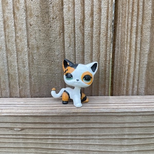 May include: A small, stylized cat figurine featuring a white base coat with patches of orange and black. The cat has large, expressive eyes with green irises, pointed ears with black tips, and a curved tail. The figure is depicted in a standing pose.