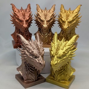 May include: A collection of dragon head sculptures in various metallic finishes. The sculptures are displayed in copper, bronze, gold, and rose gold. Each dragon head is intricately detailed with scales and a fierce expression, mounted on a square base.