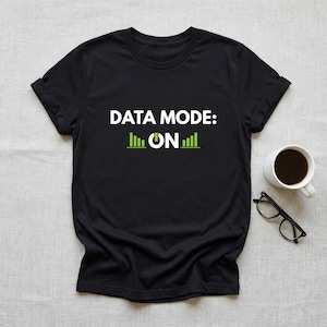 May include: A black short-sleeved t-shirt featuring the white text "DATA MODE:" above a green power symbol and the word "ON" flanked by two green bar graph icons. The shirt is displayed alongside a white coffee mug and black-rimmed glasses.