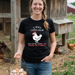 May include: Black t-shirt with the text "I MAY LOOK CALM BUT IN MY HEAD I'VE PECKED YOU 3 TIMES..." and a cartoon chicken graphic. The woman is holding a basket of eggs.