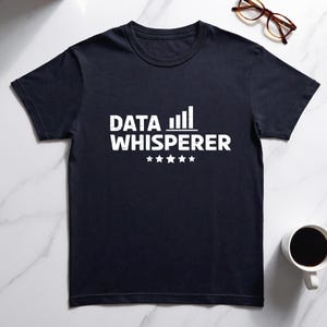 May include: A dark navy blue short-sleeved t-shirt featuring the white text "DATA WHISPERER" printed on the chest. Above the text is a small bar graph icon, and below the text are five small white stars arranged in a horizontal row.