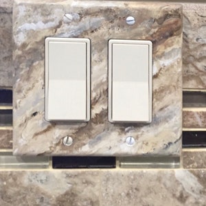 May include: Two white light switches mounted on a decorative brown and beige tile with a marble pattern.