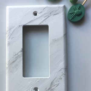May include: A white light switch cover with a marble pattern. The cover has two screw holes and a rectangular opening for the switch.
