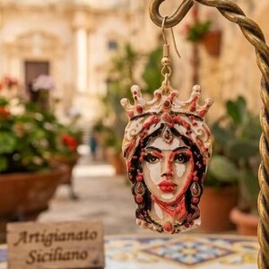 Può includere: Un orecchino pendente che raffigura il volto scolpito e dipinto a mano di una persona con corona, decorato con accenti rossi, neri e bianchi. Il pezzo è sospeso a un gancio in metallo dorato e mostra tratti del viso e gioielli dettagliati.