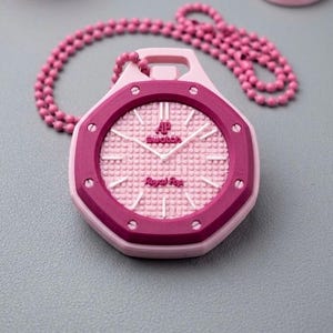 Könnte beinhalten: Eine rosa Anhängerkette mit einem Uhrendesign. Das Zifferblatt der Uhr ist hellrosa mit einem strukturierten Muster und weißen Zeigern. Die Lünette ist dunkler rosa, und der Text "AP swatch Royal Pop" ist sichtbar. Die Kette besteht aus kleinen rosa Perlen.