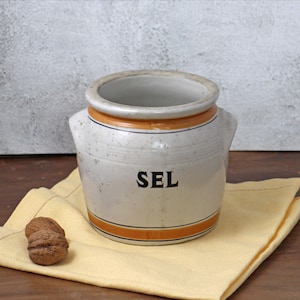 May include: A vintage ceramic salt jar with the word "SEL" in black lettering. The jar is off-white with orange and blue stripes. It has two small handles and sits on a yellow cloth next to a walnut.
