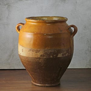 May include: A large, antique ceramic jar with a two-tone brown and tan glaze. The jar has two small handles and a wide opening. The glaze has a distressed appearance, with areas of wear and chipping, adding to its vintage charm.