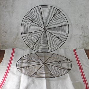 Può includere: Due griglie di raffreddamento circolari in filo metallico con un motivo a spirale concentrica. Ogni griglia è realizzata in sottile filo metallico scuro con raggi di supporto e piccoli piedini ad anello per raffreddare i prodotti da forno.