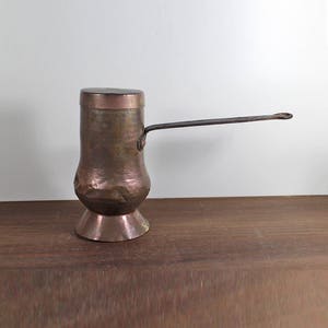 May include: A copper coffee pot with a long, thin handle and a lid. The pot has a bulbous body and a flared base. The metal has a hammered texture and a dark, aged patina. The handle is attached to the side of the pot.