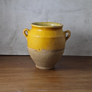 May include: A rustic, two-handled ceramic pot with a yellow upper section and a tan lower section. The pot has a rounded body and a slightly flared rim. The pot is sitting on a wooden surface.