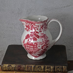 Peut inclure: Une cruche en céramique blanche avec un motif floral et paysager rouge. La cruche a une anse incurvée et un bord festonné. Le motif comprend des bâtiments, des arbres et des fleurs. La cruche est posée sur une pile de livres.