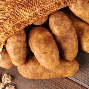 May include: Close-up of russet potatoes spilling from a burlap sack onto a wooden surface. The potatoes are oblong with brown skin. The burlap sack is a light brown color.