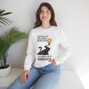 May include: White long-sleeve sweatshirt with the text "The road to brilliance begins with the admission of ignorance." The design includes a road leading to a lightbulb. The model is wearing blue jeans.
