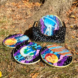 May include: Set of four round coasters with a space theme, featuring planets and galaxies in vibrant colors. The coasters are displayed on grass with a black holder with white speckles. The coasters are approximately 4 inches in diameter.