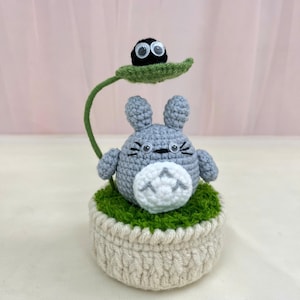 May include: Crocheted Totoro figure with a soot sprite on a leaf. The gray Totoro has a white belly and sits atop a green mossy base within a cream-colored pot. A green stem supports the leaf and soot sprite.