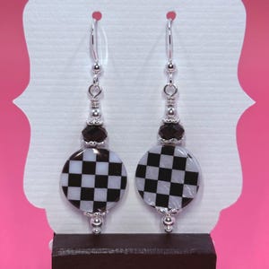 May include: A pair of dangle earrings featuring a black and white checkered design. The earrings have a silver-tone metal construction with black beads and a round checkered pendant. The earrings are displayed on a white card with a pink background.