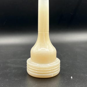 May include: A light beige bottle stopper with a cylindrical top and a flared base. The base has several horizontal ridges. The stopper is made of a translucent material and is set against a black background.