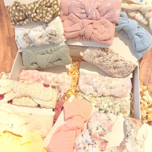 May include: A collection of assorted fabric headbands featuring various patterns including floral prints, polka dots, gingham checks, and solid colours. Each headband is tied in a knot at the centre and mounted on a white card, presented together in a box.