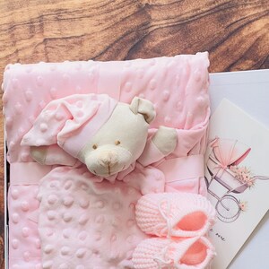 Op de afbeelding: Een babycadeauset in een witte doos, met een roze deken met een gestippelde textuur, een pluchen konijn, een bijpassende outfit en gebreide babyslofjes. Een wenskaart met een illustratie van een kinderwagen is ook inbegrepen.