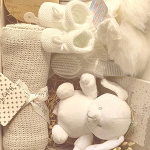 May include: A white baby gift set in a box, including a plush bunny, knitted booties, a blanket, a brush, and a wooden sign that says "Hello World". The set is decorated with a ribbon and star-shaped tag.