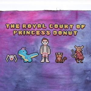 May include: Cross-stitch art on purple fabric with the text "THE ROYAL COURT OF PRINCESS DONUT." The design features pixelated characters including a person, a dinosaur, a cat, and a rabbit. The background has a tie-dye effect.