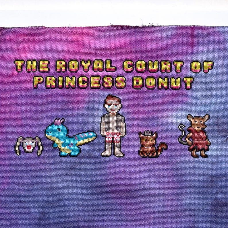May include: Cross-stitch art on purple fabric with the text "THE ROYAL COURT OF PRINCESS DONUT." The design features pixelated characters including a person, a dinosaur, a cat, and a rabbit. The background has a tie-dye effect.