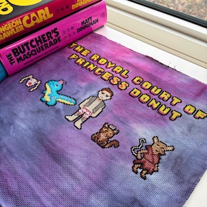 May include: A cross-stitch piece with the text "THE ROYAL COURT OF PRINCESS DONUT" in yellow. The piece features pixelated characters and a purple and blue tie-dye background. Books are in the background.