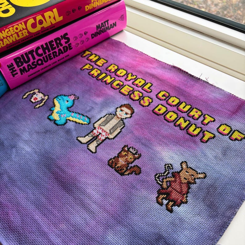 May include: A cross-stitch piece with the text "THE ROYAL COURT OF PRINCESS DONUT" in yellow. The piece features pixelated characters and a purple and blue tie-dye background. Books are in the background.