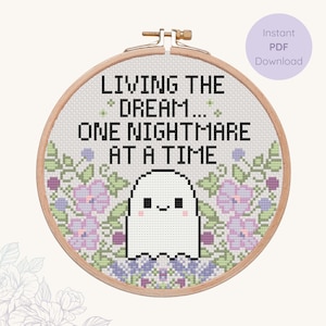 May include: Cross-stitch embroidery in a wooden hoop with the text "LIVING THE DREAM... ONE NIGHTMARE AT A TIME." A cute ghost is centered below the text, surrounded by floral designs in purple and green. A circular label reads "Instant PDF Download."