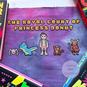 May include: A cross-stitch design on purple fabric features the text "THE ROYAL COURT OF PRINCESS DONUT" in yellow and orange. Pixelated characters include a man, a dinosaur, and other creatures. The design is surrounded by books.