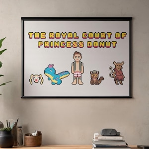 May include: Framed cross-stitch art featuring the text "THE ROYAL COURT OF PRINCESS DONUT" above pixelated characters. The characters include a bunny, a blue dinosaur, a person, a cat, and a rodent. The artwork is displayed on a wall.
