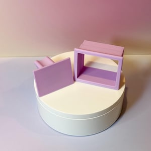 May include: A lilac-colored, rectangular frame and a matching rectangular piece are displayed on a white, circular platform. The background is a gradient of pink and purple.