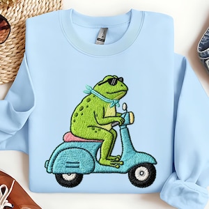 Könnte beinhalten: Hellblaues Sweatshirt mit einem gestickten Design einer grünen Frosch mit Sonnenbrille und blauem Schal, der einen türkisfarbenen Roller fährt. Der Frosch sitzt auf einem rosa Sitz. Der Roller hat schwarze Räder.
