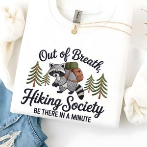 May include: White sweatshirt with the embroidered text "Out of Breath, Hiking Society, Be There In A Minute." The design features a raccoon with a backpack and two green pine trees. A gold necklace is draped across the sweatshirt.