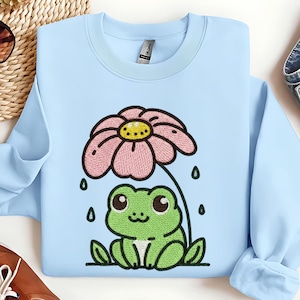 May include: Light blue sweatshirt featuring an embroidered design of a green frog under a pink flower, with raindrops. The frog has large eyes and a smiling expression. The flower has a yellow center. The sweatshirt has a crew neck.