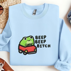 May include: Light blue sweatshirt with an embroidered design of a green frog driving a red car. The text "BEEP BEEP BITCH" is embroidered in black on the right side of the frog.