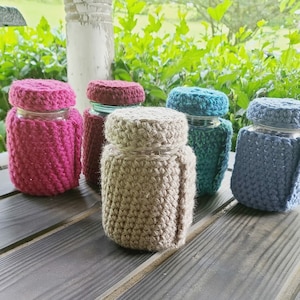 May include: Five glass jars with crocheted covers in various colors, including pink, brown, teal, and blue. The jars are sitting on a wooden surface.