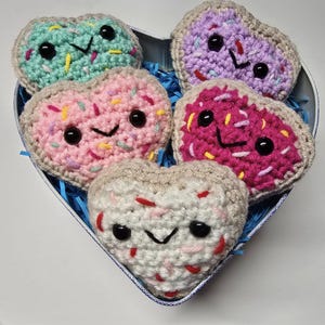 Può includere: Cinque fette di pane all'uncinetto a forma di cuore con diverse glassature e codette colorate. Le fette di pane sono in una scatola a forma di cuore con carta velina blu e bianca.