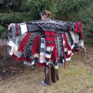 May include: A long, multi-coloured knitted cardigan with a patchwork design. The cardigan is made from different shades of grey, white, and red. The cardigan has a frayed edge and is being worn by a person.