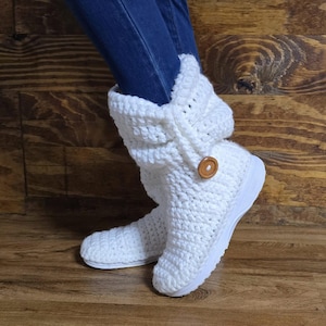 以下が含まれることがあります： 茶色のボタンが付いた白いクロシェのゆったりとしたブーツ。ブーツは白いソールで、木製の床の上で履かれています。