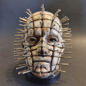 May include: A white ceramic sculpture of a human head with a grid pattern and numerous metal pins protruding from the surface. The sculpture is mounted on a black base.