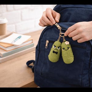 Könnte beinhalten: Zwei grüne, gehäkelte Gurken-Schlüsselanhänger mit gestickten Gesichtern und goldfarbenen Kettenanhängern. Eine Gurke hat ein wütendes Gesicht und eine Fliege, die andere ein lächelndes Gesicht und eine Blume. Die Schlüsselanhänger sind an einem marineblauen Rucksack befestigt.