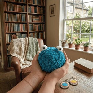 Peut inclure: Une boule bleue crochetée avec de grands yeux blancs et noirs est tenue dans les deux mains. L'arrière-plan comprend une table en bois, une fenêtre avec des plantes en pot et une bibliothèque remplie de livres. Deux petits pendentifs sont sur la table.