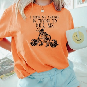 May include: Orange crew neck sweatshirt with the text "I THINK MY TRAINER IS TRYING TO KILL ME" above a black skeleton lifting weights. A white mug with a yellow smiley face is held in the hand.
