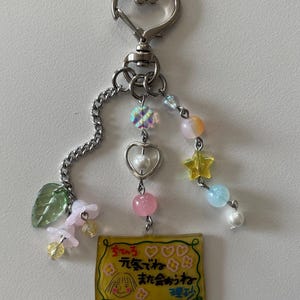 May include: A silver-toned keychain with a heart-shaped clasp and a chain. The keychain features a yellow rectangular charm with a cartoon drawing and Japanese text, along with colorful beads and charms in pink, yellow, and blue.