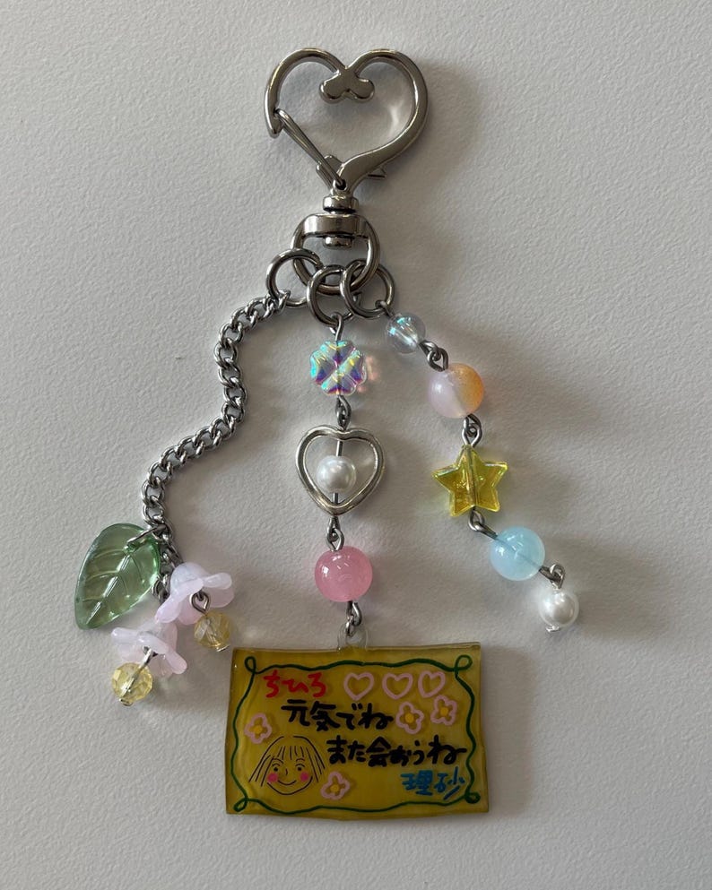 May include: A silver-toned keychain with a heart-shaped clasp and a chain. The keychain features a yellow rectangular charm with a cartoon drawing and Japanese text, along with colorful beads and charms in pink, yellow, and blue.
