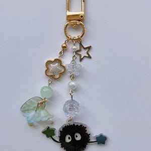 May include: A gold-colored keychain with various charms. Charms include a gold flower, a clear star, a white pearl, a blue flower, and a black soot sprite with white eyes. The keychain is attached to a gold clasp.